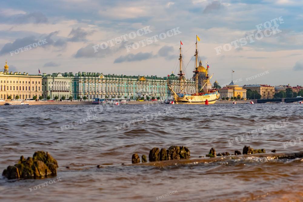 На фотографии представлен вид на Зимний дворец и реку Неву в Санкт-Петербурге с набережной. В центре кадра находится парусник с поднятыми парусами, который является репликой исторического корабля и придает изображению нотку величия и историзма. Вода реки Невы бурлит, на переднем плане видны камни набережной, волнение воды указывает на ветреную погоду. Это фото может быть использовано для иллюстрирования статей о морской истории России, туристических брошюр, образовательных материалов по истории Петербурга или как элемент декора, отражающий культурное наследие города.